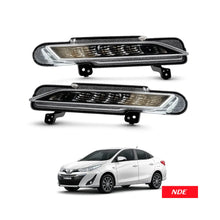 A pair of automotive fog lights shown above a white sedan on a light gray background. The fog lights have black housings, clear outer lenses, and a smoked amber center section.