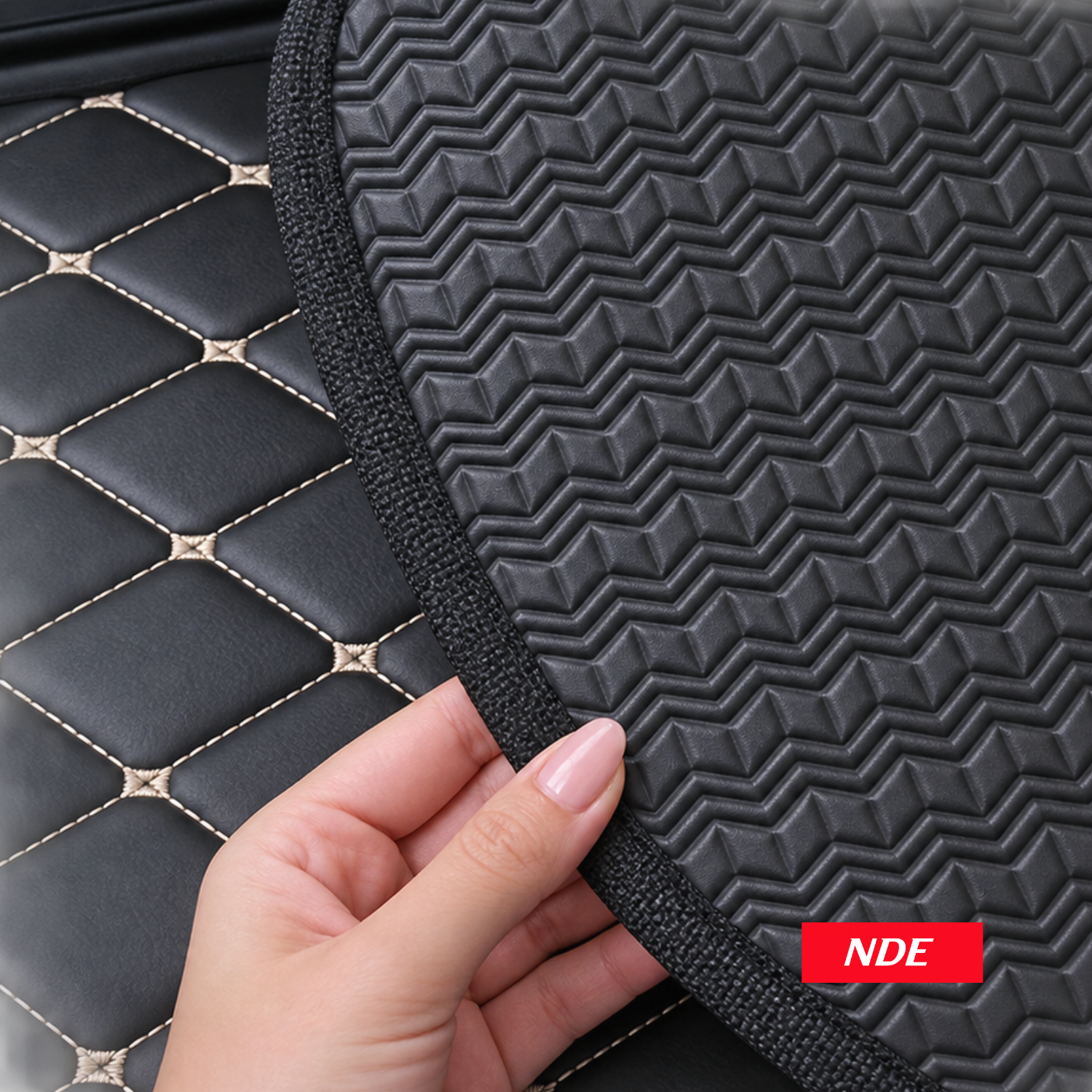 A close-up of a black textured car seat pad with a geometric chevron pattern, held by a hand over a black quilted leather seat with gold stitching. A red badge with the text 'NDE' is visible in the bottom right corner.
